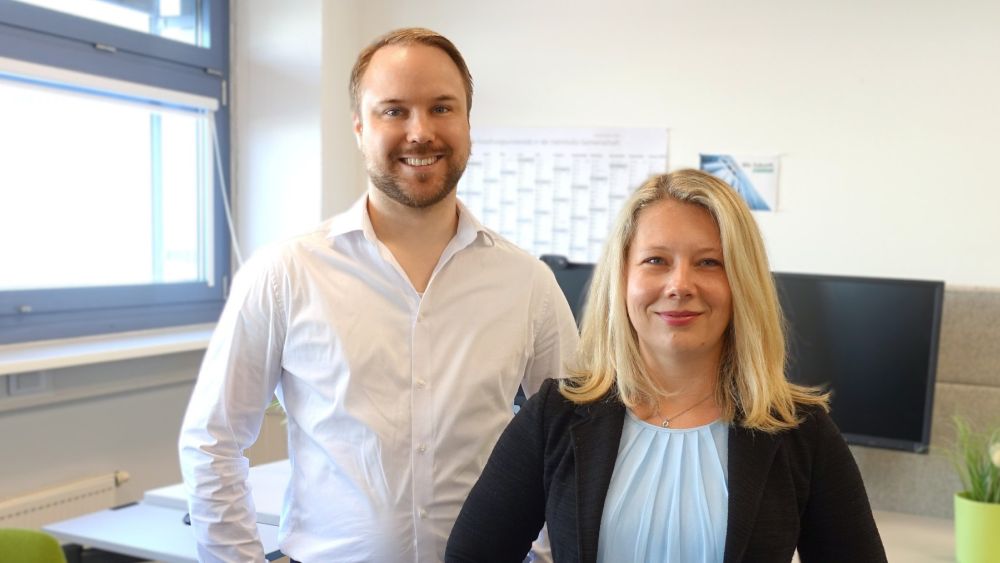Zwei Personen in einem Büro, im Hintergrund ein Schreibtisch mit PC-Ausstattung