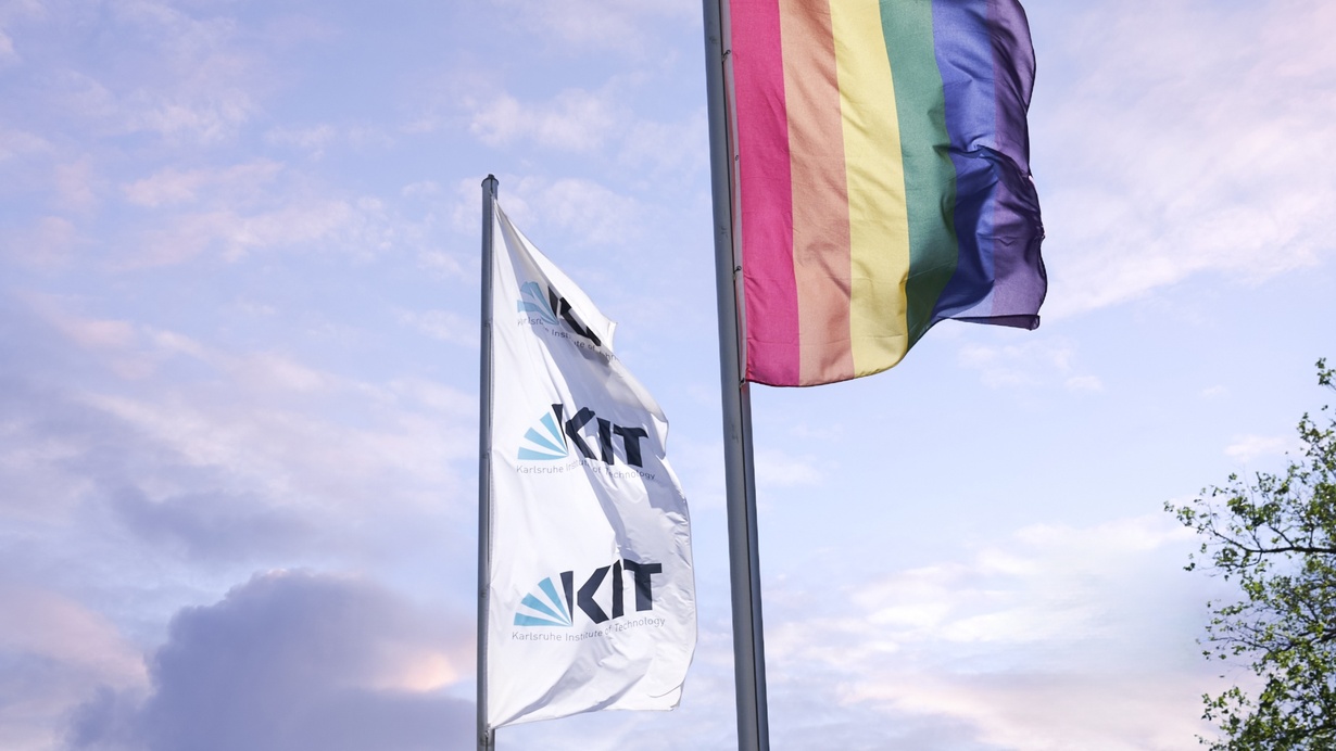 Two flags - one white with KIT logo, the other a rainbow flag - flying against a blue sky.